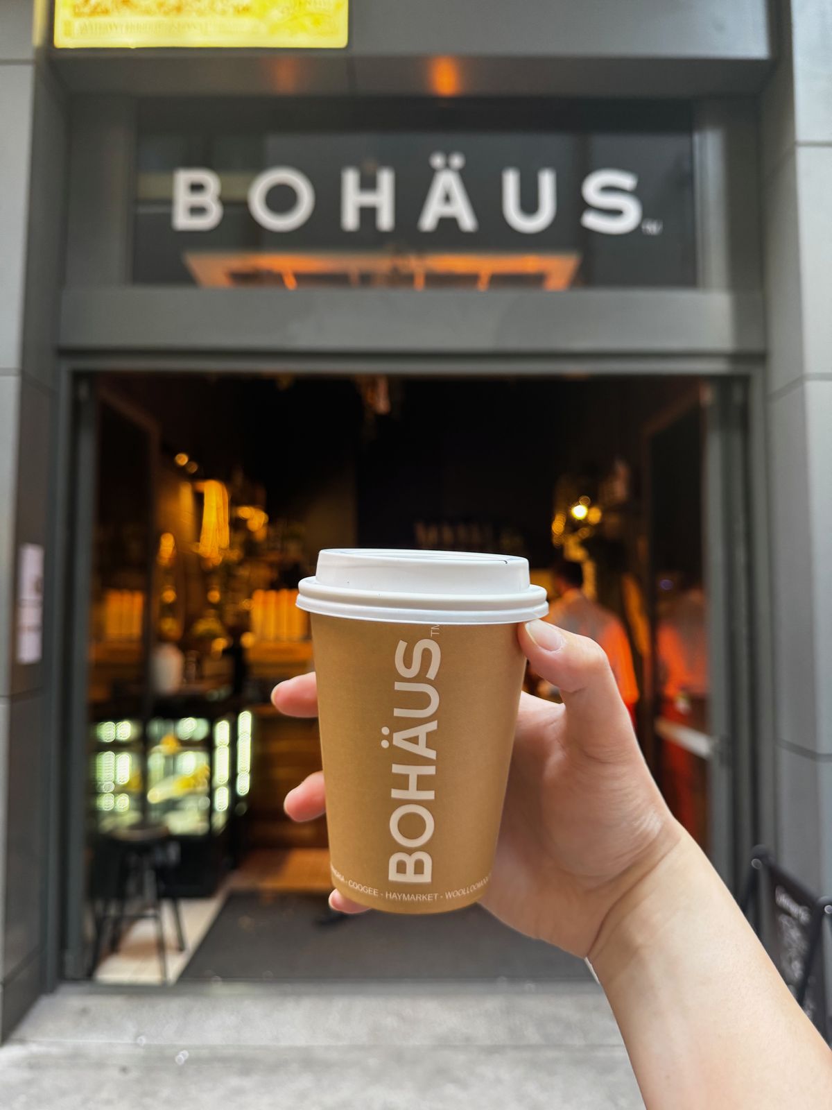 Takeaway coffee cup branded Bohaus held in front of the cafe entrance