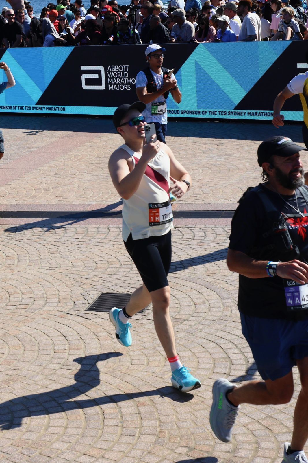 Runner in a white and maroon singlet filming on a phone while running through the finish area