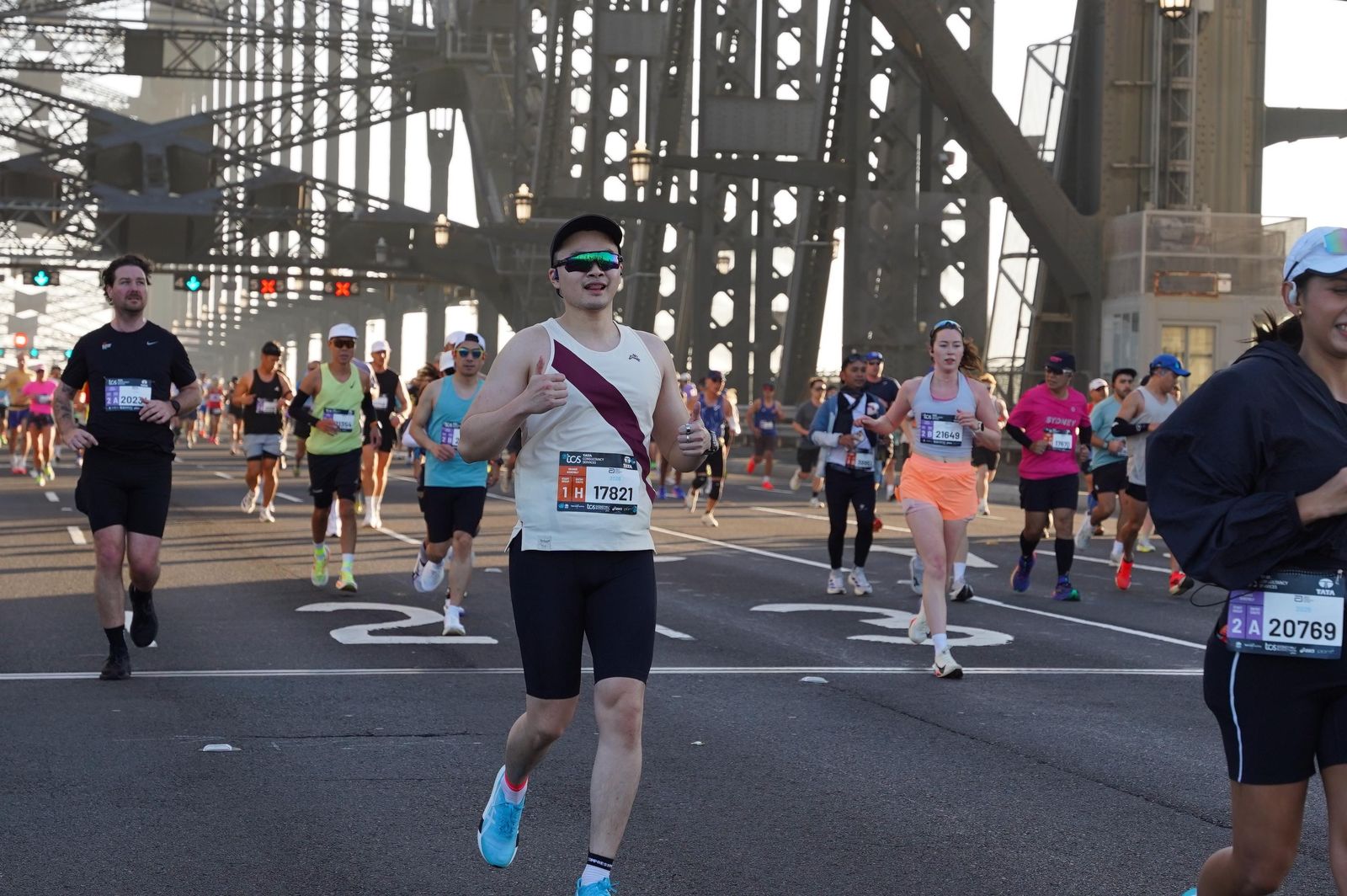 Runner in a white and maroon singlet giving a thumbs up while crossing the Sydney Harbour Bridge among other marathon runners